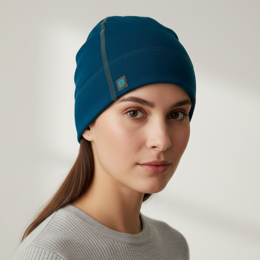 A woman wearing a peacock merino wool beanie hat and a gray ribbed knit top, photographed against a soft light gray background.