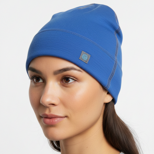 A woman wearing a bright blue merino wool beanie hat with a small square logo patch on the side, photographed against a clean light gray studio background.